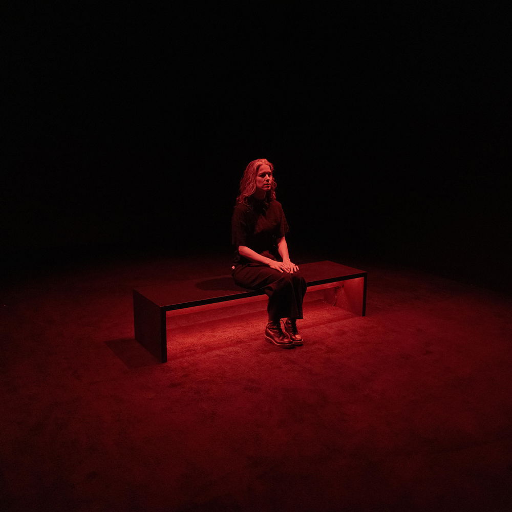 A woman wearing a black top, black pants, and black boots sits on a bench in the middle of an empty, dark room. She is lit from above by soft red light.