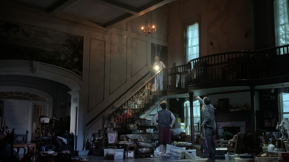 Two men stand on a stage made up to look like a grand mansion room with a central staircase and balcony. It appears to be night and the room is disheveled, filled with boxes, baskets, bins, and other objects. They look up to the top of the staircase where a third man in pajamas shines a flashlight down at them.